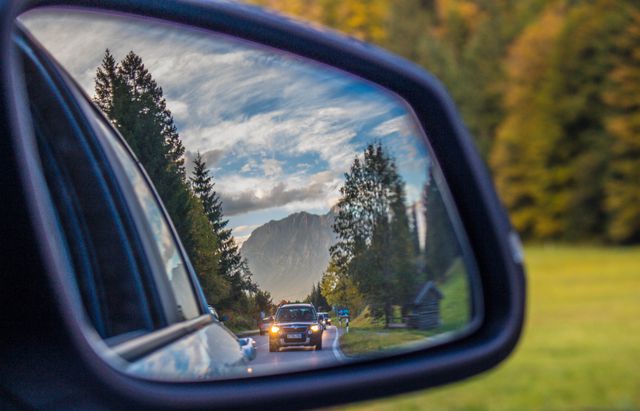 Reflection of a stunning mountain landscape in a car's rearview mirror features a tranquil road with trees and a cloudy sky. Ideal for use in travel blogs, automotive promotions, adventure-themed advertisements, and scenic compositions depicting journeys.