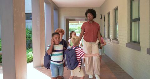African american teacher leading diverse kids down school hallway with backpacks