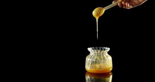 Golden honey drizzling from a wooden dipper into a decorative glass jar against a black background. This image highlights the rich, viscous texture and natural golden hue of fresh honey. Perfect for use in advertisements, food blogs, health and wellness articles, or as a visual representation of natural sweeteners.