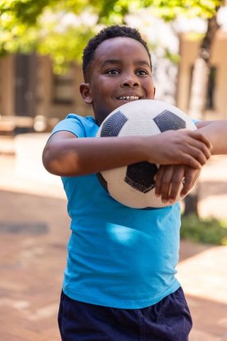 Joyful african american boy holding soccer ball in sunlit urban courtyard