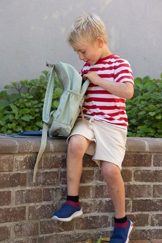 Blond boy sitting on brick ledge unzipping light green backpack with tablet outdoors