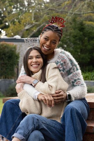 Female couple hugging in garden wearing merry christmas headbands, cozy knitwear holiday