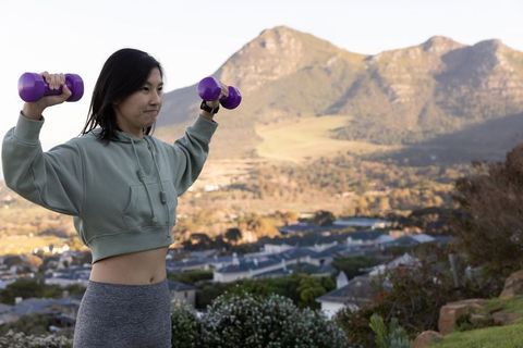 Asian woman lifting purple dumbbells during outdoor workout on hilltop with mountain view
