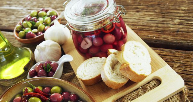 Rustic display featuring a variety of Mediterranean olives, pickled vegetables in jar, white garlic bulbs, fresh bread slices on wooden table. Ideal for illustrating healthy eating, Mediterranean cuisine, gourmet food advertisements.