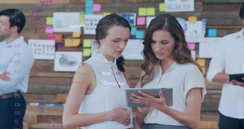 Two women reviewing tablet with code overlay and sticky notes in creative teamwork scene