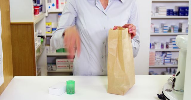 Pharmacist at the counter inside a pharmacy packing a prescription into a paper bag. Ideal for illustrating themes related to healthcare, medicine dispensation, customer service in retail pharmacies, and the role of pharmacists in ensuring correct medication distribution.