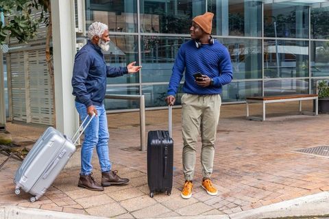 African american men talking by urban glass building pulling suitcases using smartphone