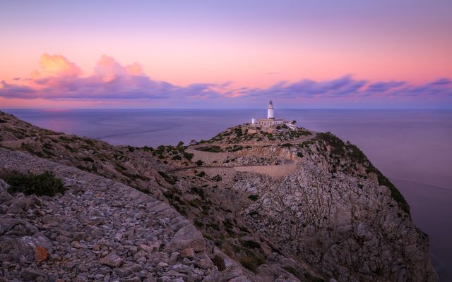 Photo showcasing a lighthouse on the edge of a rugged cliff during a vibrant sunset. The sky transitions from pink to purple with clouds creating an eye-catching scene. Ideal for use in travel blogs, brochures, nature magazines, and websites related to tourism and coastal vacations.