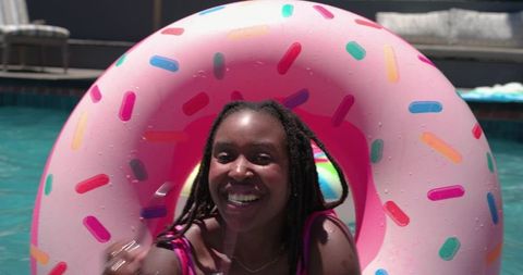 Smiling african american woman floating on pink donut pool float enjoying sunny backyard