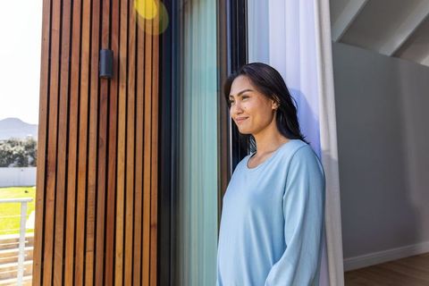 Asian woman standing at sliding door holding curtain enjoying sunlit garden view