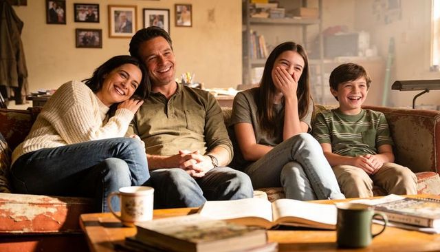 Laughing family relaxing on worn sofa with open book, mugs and warm natural light