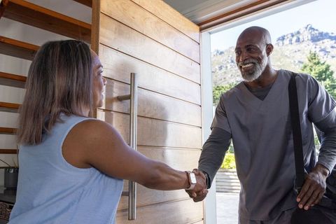 Smiling african american nurse shaking hands with mature woman at home entrance