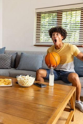 Young man cheering with basketball on sofa, popcorn and drink on coffee table during game