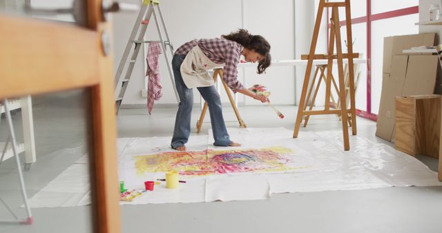This image shows a female artist engaged in creating an abstract painting on a large canvas placed on the floor of a modern studio. It can be used to evoke themes of creativity, contemporary art, and the artistic process. Ideal for articles, websites, and promotions related to art, workshops, creativity-enhancing environments, and modern studios.
