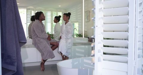 African american couple relaxing on vanity in sunlit modern bathroom wearing robes