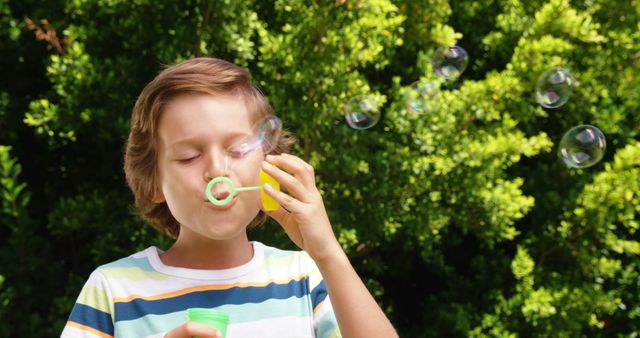Young boy blowing soap bubbles outdoors on a sunny day. Perfect for themes related to childhood, play, happiness, outdoor activities, summer fun, and family life. Suitable for advertising, editorial content, and parenting articles.