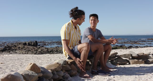 Two young adult friends casually chatting while sitting on a rock wall at the beach. They appear relaxed and at ease, enjoying a sunny day by the ocean. Ideal for use in lifestyle, leisure, and friendship-themed projects, summer vacation advertisements, or social media content promoting outdoor activities and bonding experiences.