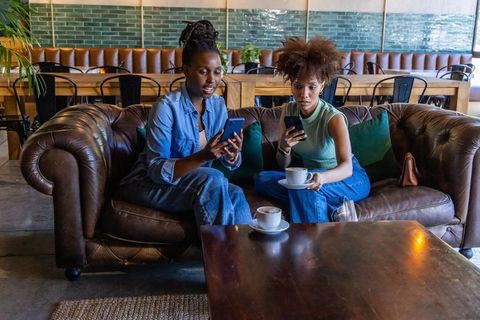 Two friends scrolling smartphones and drinking coffee on leather sofa in modern urban cafe