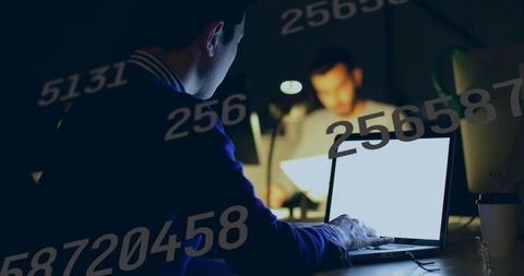 Asian man typing on laptop late in dim coworking office with numeric overlay, coding analytics