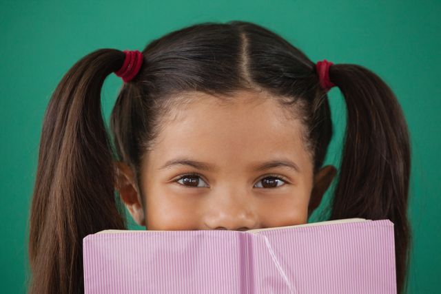 This image shows a young schoolgirl with pigtails hiding behind a book against a green background. It can be used for educational materials, school promotions, children's book covers, and articles about childhood learning and education.
