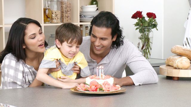 Family gathered in kitchen with young child blowing out birthday candles on a beautifully decorated cake. Ideal for concepts of family celebrations, joy, childhood moments, and special occasions.