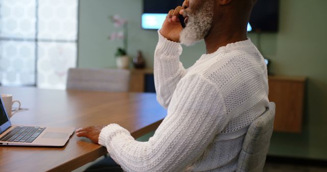 Older man with white beard on phone working at laptop in home office - Download Free Stock Images Pikwizard.com