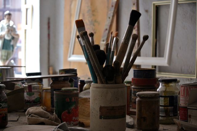 Various brushes in a ceramic holder surrounded by jars of paint and other materials on cluttered table, suggesting active workspace for an artist. Ideal for depicting creativity, artisanal processes, artistic ambiance or the dynamic environment of art studios. Can be used for art supply promotions, creativity blogs or as a background for artistic content.