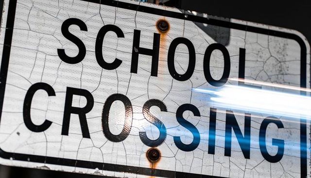 Weathered school crossing sign with cracked reflective sheeting, rusted bolts and light streak