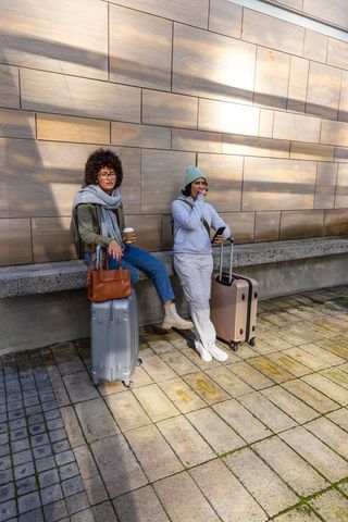 Two women waiting with rolling luggage, coffee and phones at sunlit urban plaza bench