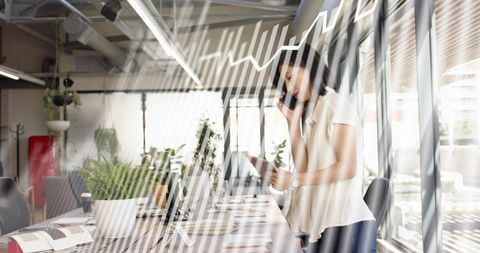 Businesswoman talking on phone holding tablet in sunlit open-plan coworking office with glass walls