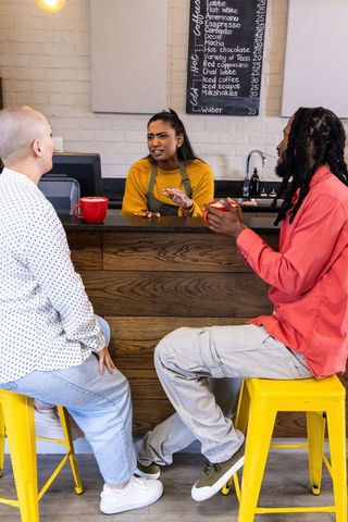 Indian barista gesturing while speaking to customers at colorful cafe counter with yellow stools