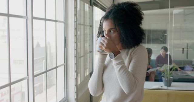 Woman standing by office window drinking coffee, taking a break from work. Stylish workplace environment with a modern interior. Useful for promoting professional settings, work-life balance content, or coffee-related products.