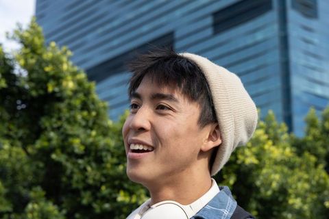 Asian teen smiling and looking up in urban park wearing beige beanie and headphones