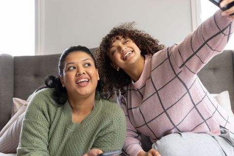 Two diverse women taking cozy bedroom selfie in knit sweaters, laughing and smiling