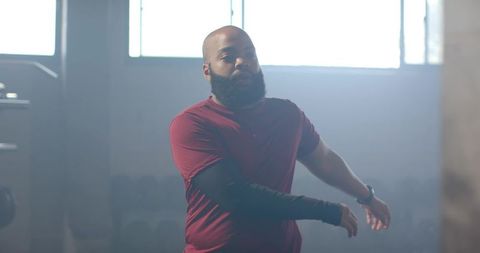 Bearded african american man warming up in gym wearing compression sleeve, backlit fitness