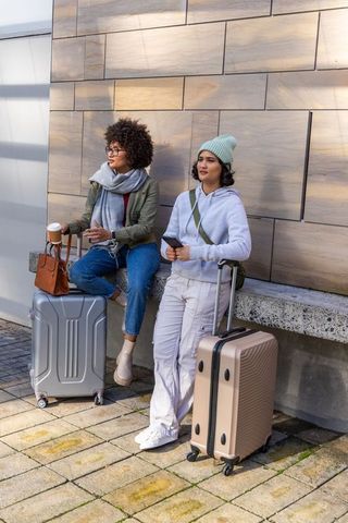 Female friends waiting with rolling suitcases outside modern building preparing for travel