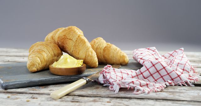 Golden croissants are paired with creamy butter on a rustic slate board, evoking a cozy, gourmet breakfast atmosphere. A red and white checkered napkin adds a touch of homey charm to the delicious scene.