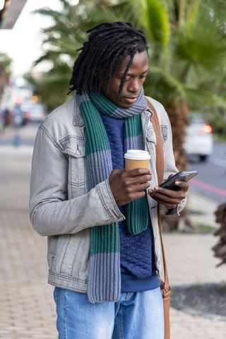 African american commuter checking smartphone while holding coffee cup on city sidewalk