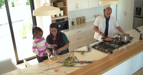 Black family cooking at sunlit kitchen island, mother guiding daughter grating cheese