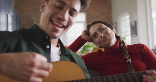 Happy biracial couple sitting on sofa in living room playing guitar. quality time, relaxing together at home.