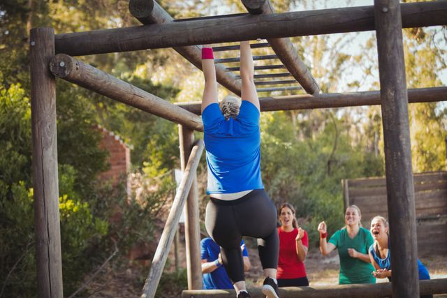 Woman climbing monkey bars while teammates cheer her on during an outdoor obstacle course training in a boot camp. Ideal for use in fitness, teamwork, motivation, and outdoor activity promotions.