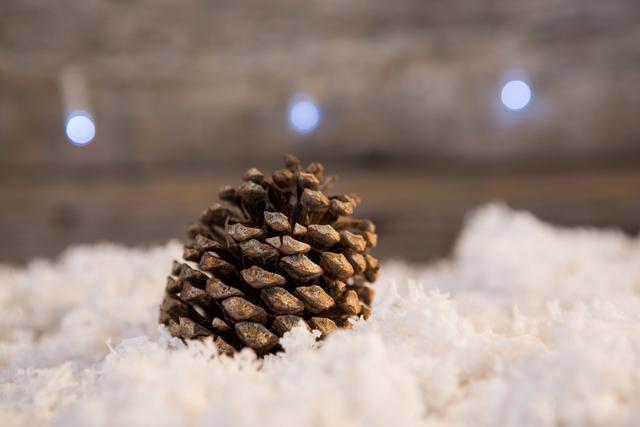 Pine cone resting on artificial snow with blurred festive lights in background. Ideal for holiday greeting cards, winter-themed decorations, and Christmas marketing materials.