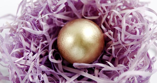 Gold Easter egg nestled in a bed of purple shredded paper creating a festive and luxurious look. Useful for Easter-themed promotions, holiday greeting cards, and springtime decoration inspirations. Suitable for blog posts about Easter traditions and DIY craft ideas.