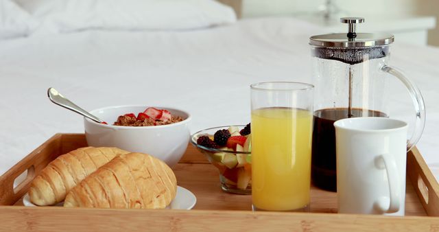 Breakfast tray featuring fresh juice, coffee press, croissants, granola with berries, and fruit salad. Perfect for depicting hotel room service, cozy morning meals in bed, or healthy breakfast lifestyles.