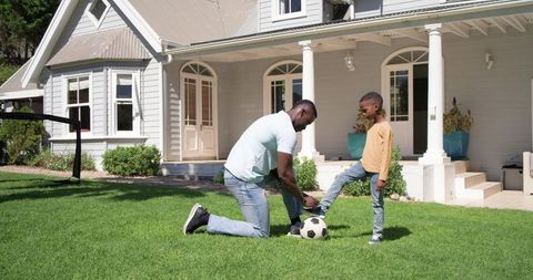 African american father tying son's shoelace while kneeling on lawn with soccer ball