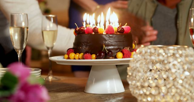 A chocolate cake adorned with lit candles and fresh berries is presented on a table, with copy space. Celebratory drinks and flowers add to the festive atmosphere, suggesting a birthday or special occasion.