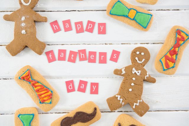 Overhead view of gingerbread cookies decorated with colorful icing, arranged around 'Happy Father's Day' text on white wooden table. Ideal for Father's Day greeting cards, social media posts, festive advertisements, and holiday-themed blog content.
