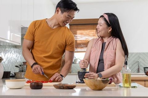 Asian couple preparing dinner in sunlit kitchen chopping tomatoes and grinding fresh herbs