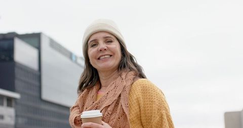 Smiling mature woman holding coffee cup wearing knit beanie and scarf on urban street