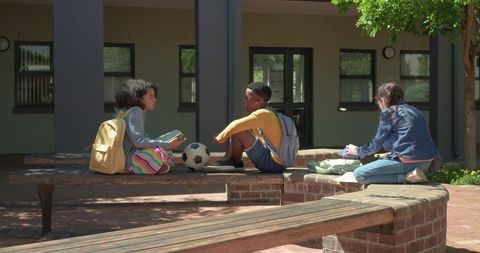 Teen friends sitting on school courtyard bench passing soccer ball near backpacks